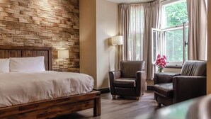 A hotel room with a wooden headboard bed, a stone wall, a large window with curtains, and two leather armchairs.