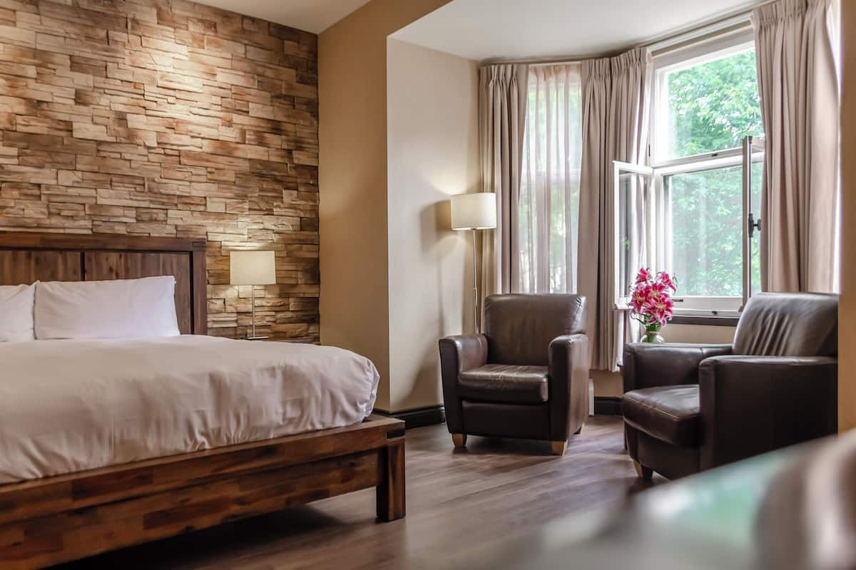 Habitación de hotel con cama de madera, cabecera de madera, pared de piedra, un gran ventanal con cortinas y dos sillones de cuero.