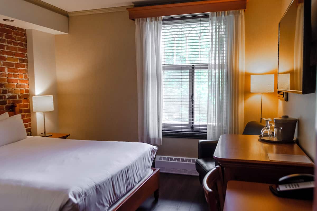 Habitación de hotel con cama, escritorio, silla, lámpara y ventana con persianas.