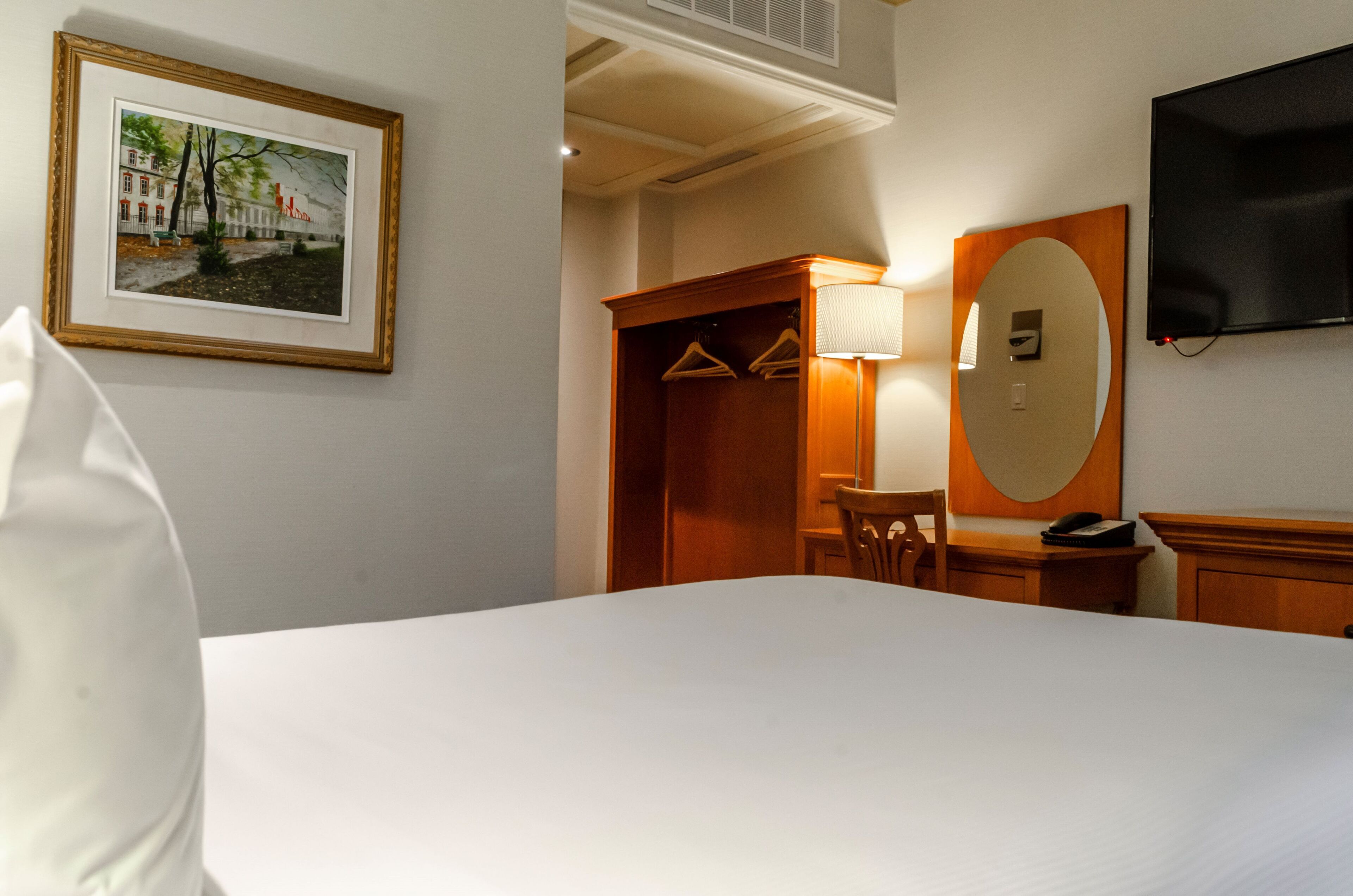 Habitación de hotel con cama, armario de madera, escritorio con silla, televisor montado en la pared y un cuadro en la pared.