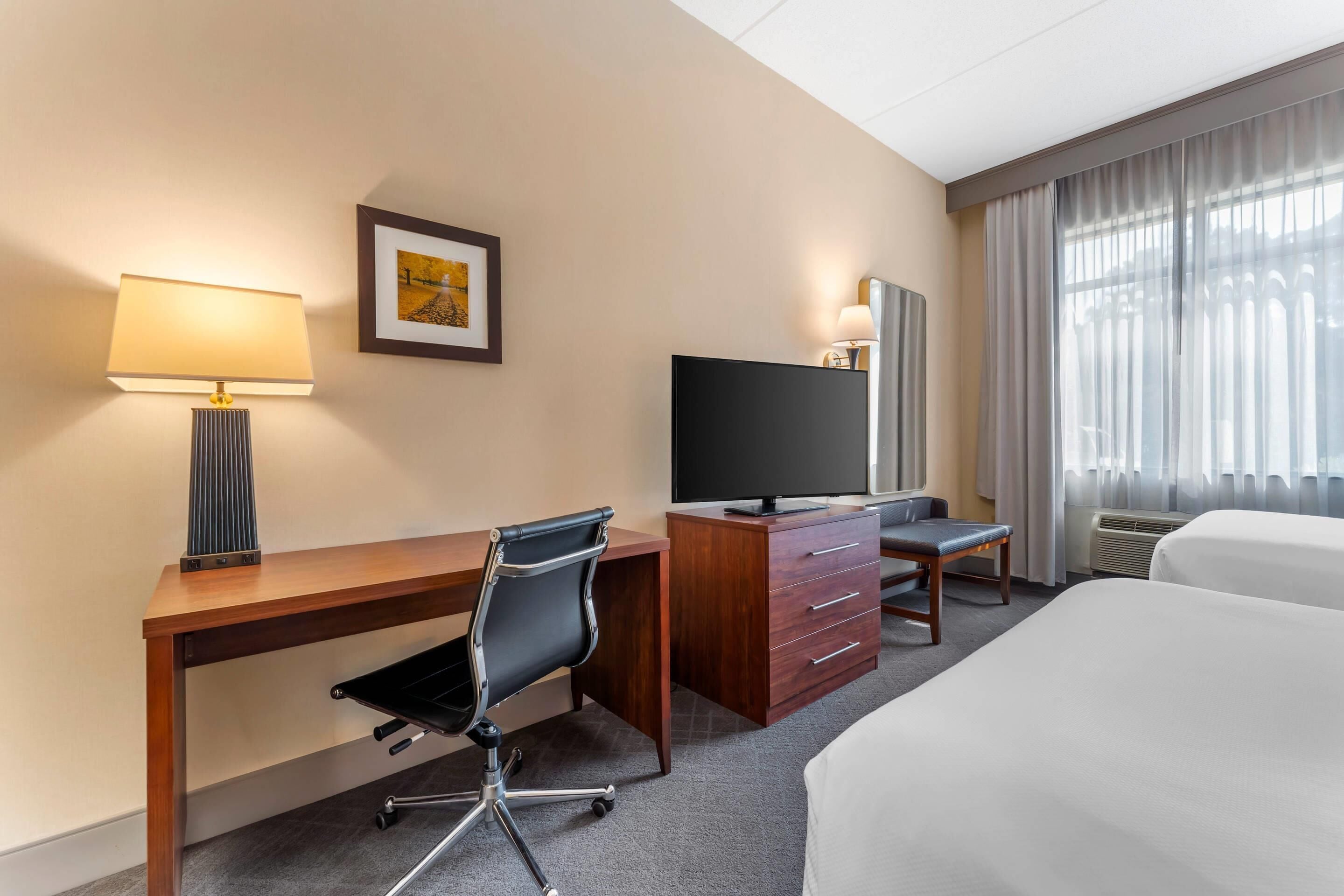 A hotel room with a wooden desk, a black office chair, a television, a bed, and a window with curtains.
