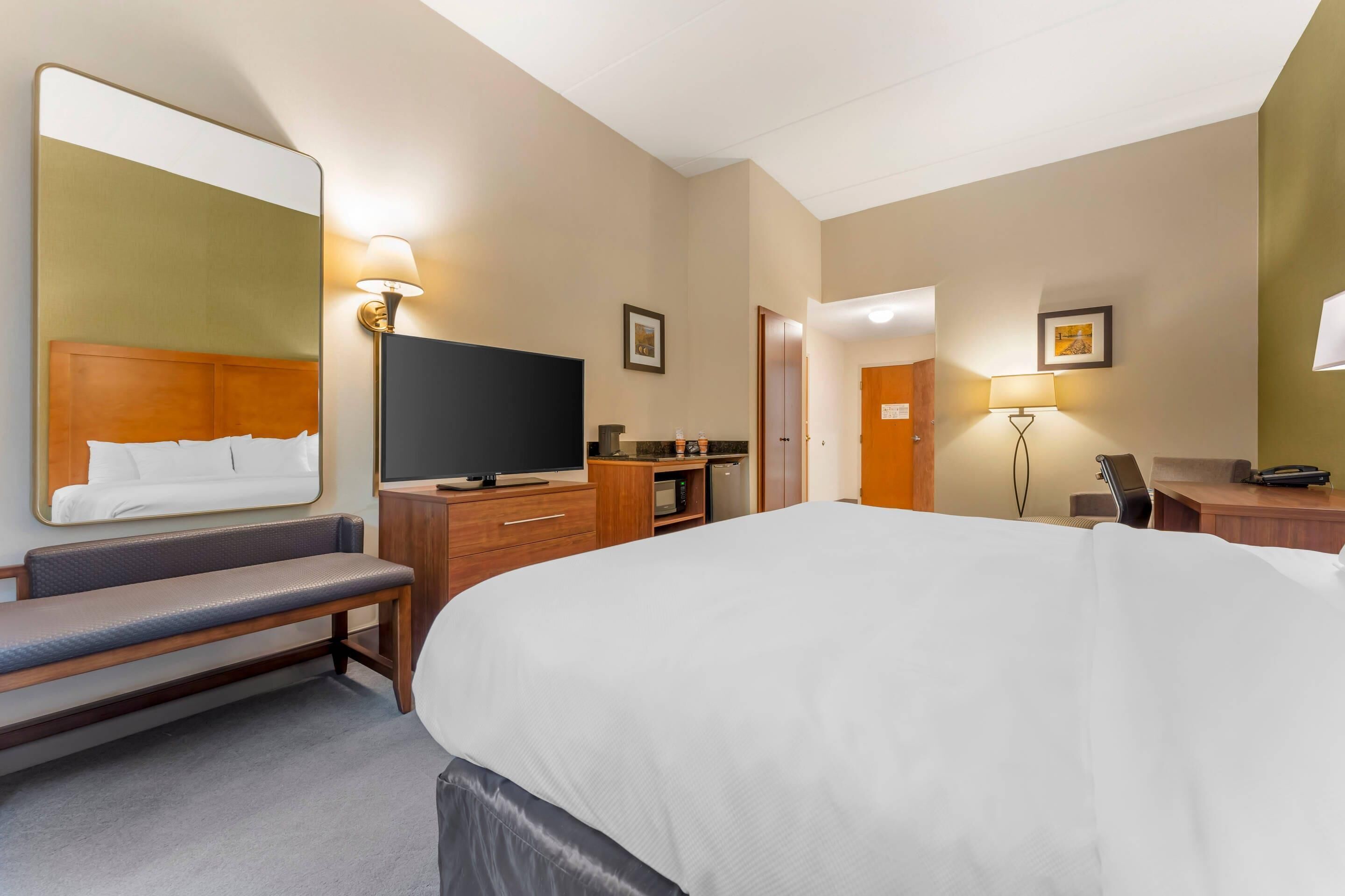 Una habitación de hotel con una cama grande, televisión, banca, escritorio y espejo.