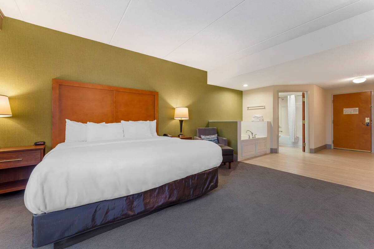Habitación de hotel con una cama grande, una mesita de noche, una lámpara, una silla y un baño visible a través de una puerta abierta.