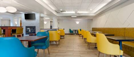 A modern restaurant with wooden flooring, yellow and blue chairs, and a flat-screen TV mounted on the wall.