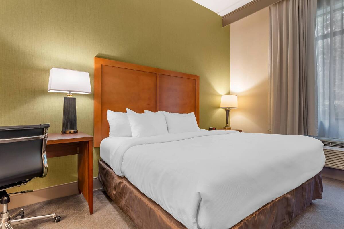 Habitación de hotel con una cama grande, un escritorio con silla, dos lámparas de noche y una ventana con cortinas.