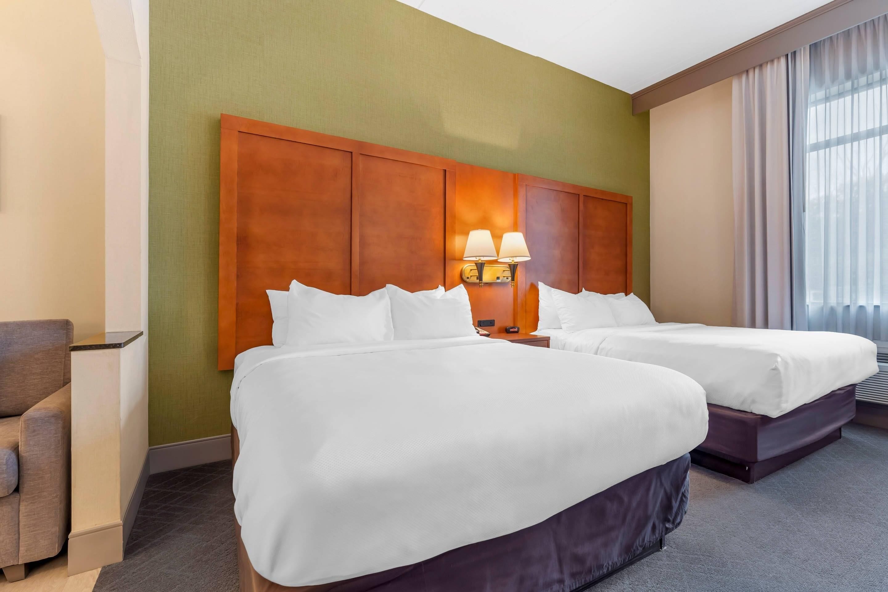 Una habitación de hotel con dos camas, una mesita de noche con lámparas, una silla y un ventanal con cortinas.