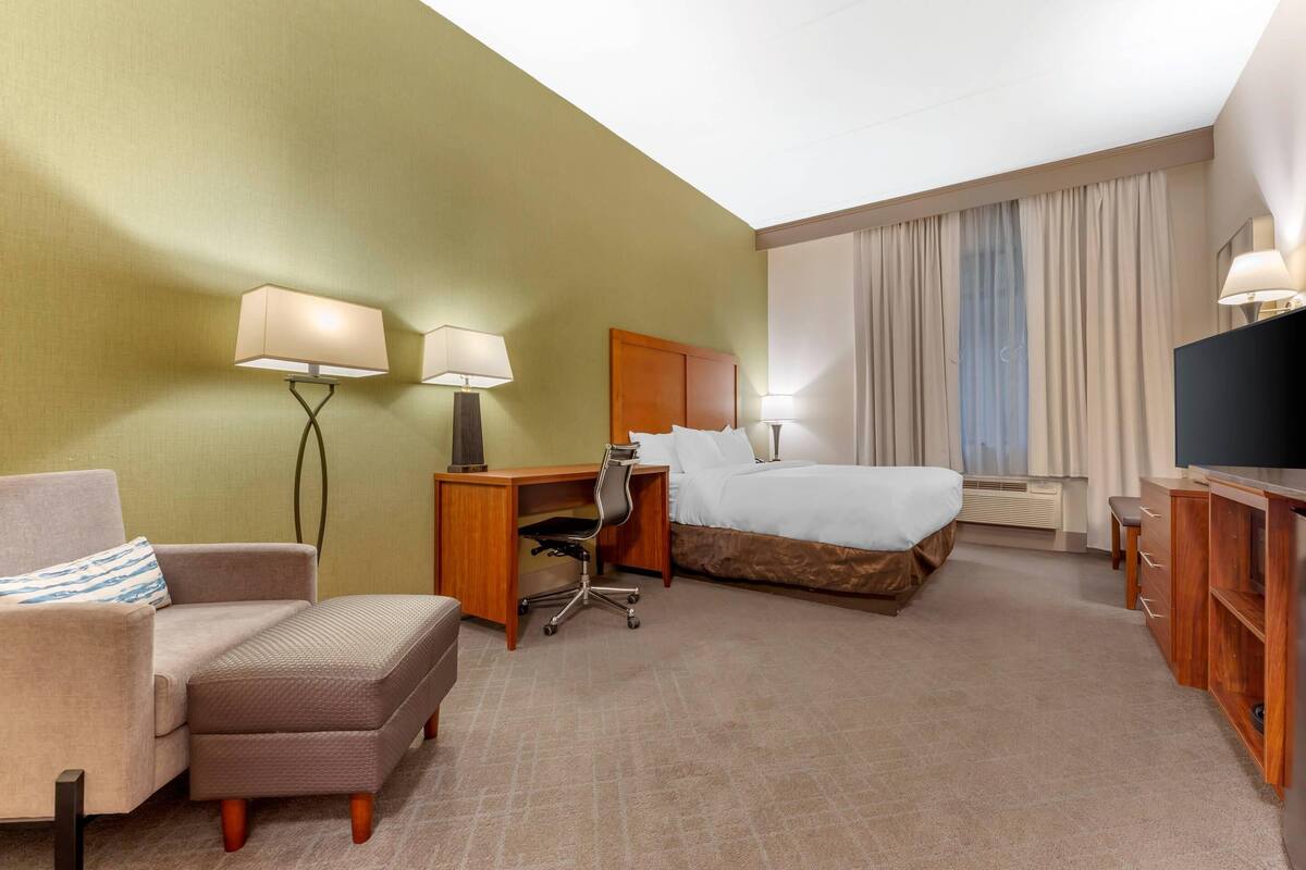 Habitación de hotel con cama, escritorio, silla, sofá, televisor y ventana con cortinas.