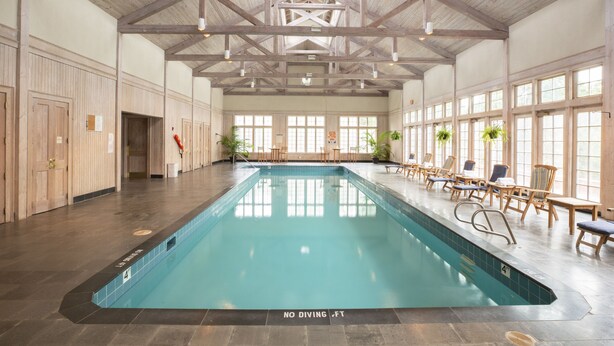 Una piscina interior con techo alto y vigas de madera, rodeada de sillones de descanso y plantas en macetas.