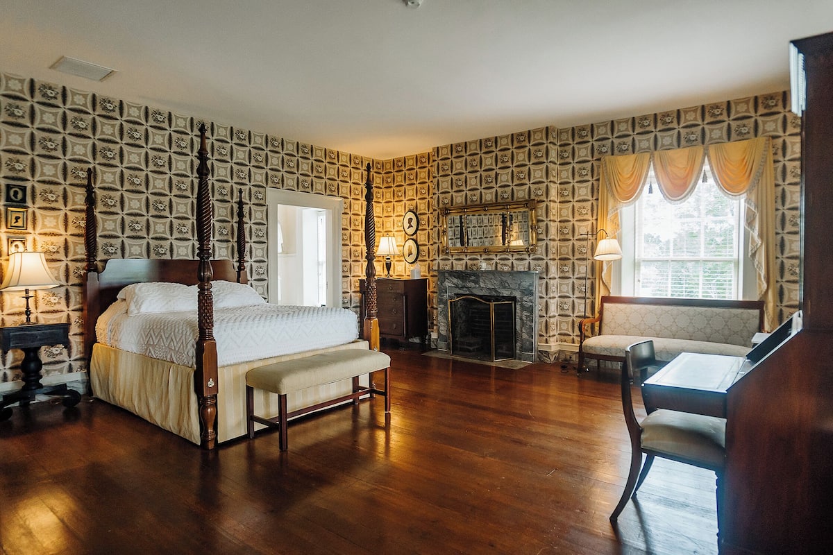 A traditional bedroom with a four-poster bed, a fireplace, and a seating area with a table and chair.