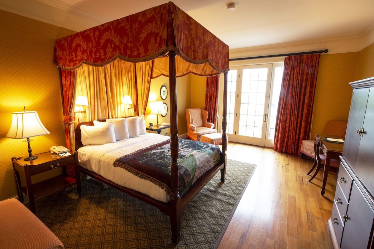 A four-poster bed with a canopy, a desk with a lamp, a chair, and a wardrobe in a hotel room.