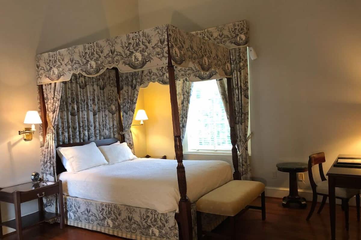 A four-poster bed with floral curtains, bedside lamps, and a small table with a chair.