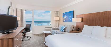 Habitación de hotel con una cama grande, un escritorio con televisor, un sofá y vistas al mar.