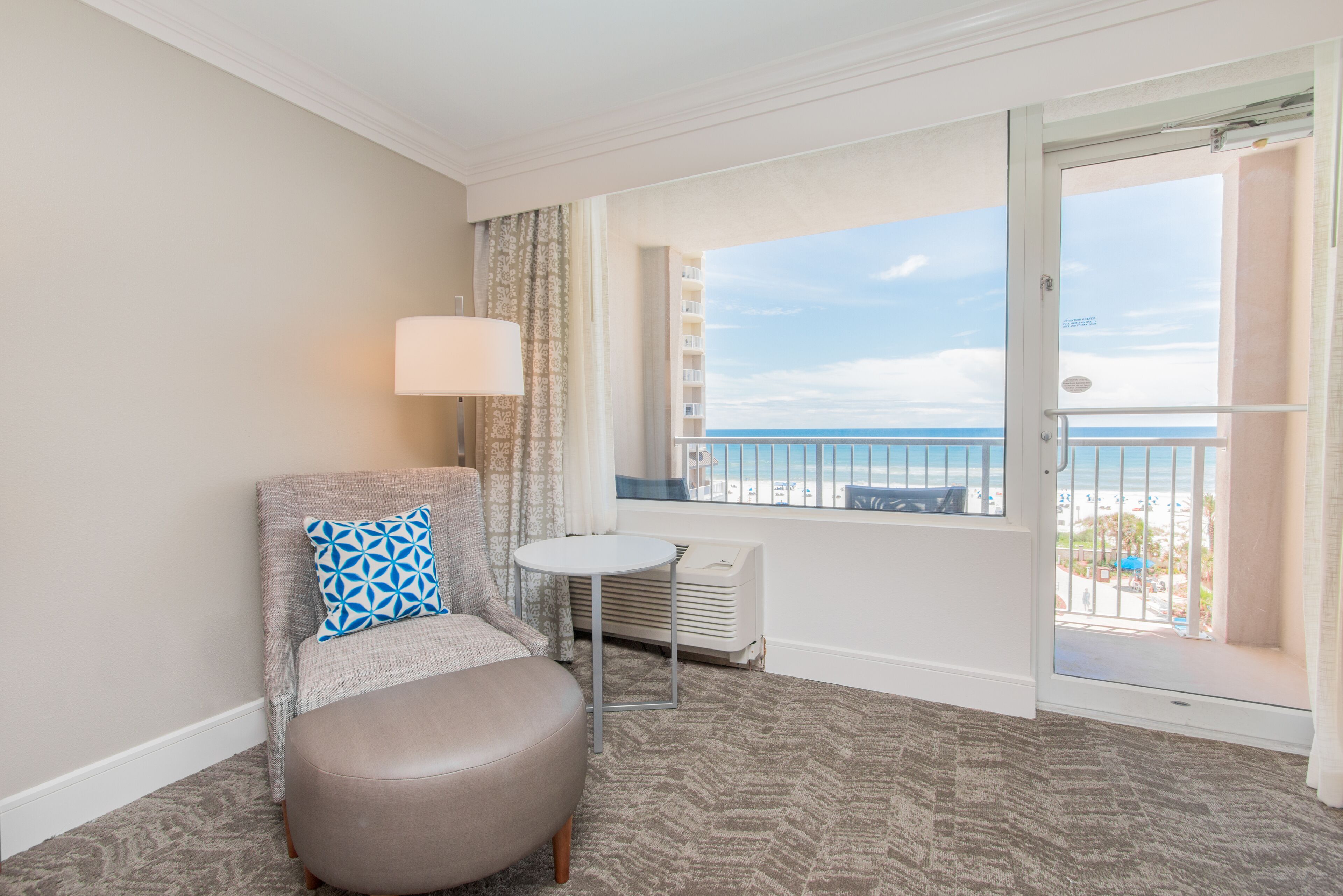 Habitación de hotel con vista a la playa, una mesa redonda, una silla con un cojín estampado y un radiador.
