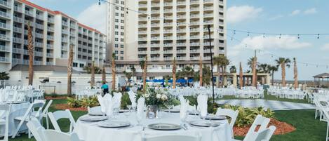 Un banquet en plein air avec des tables et des chaises blanches, un grand bâtiment en arrière-plan et des palmiers.