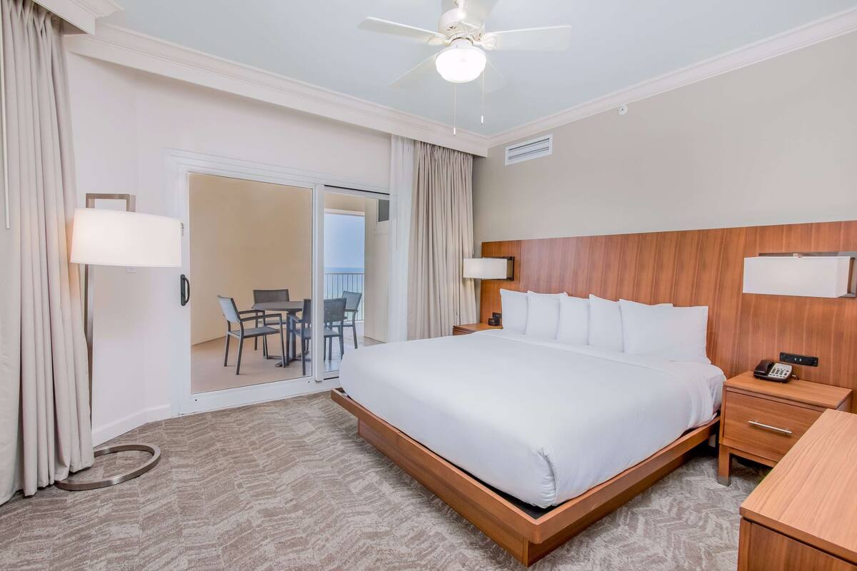 Une chambre d’hôtel avec un grand lit, des tables de chevet, un ventilateur de plafond et une vue sur un balcon doté d’un coin repas.