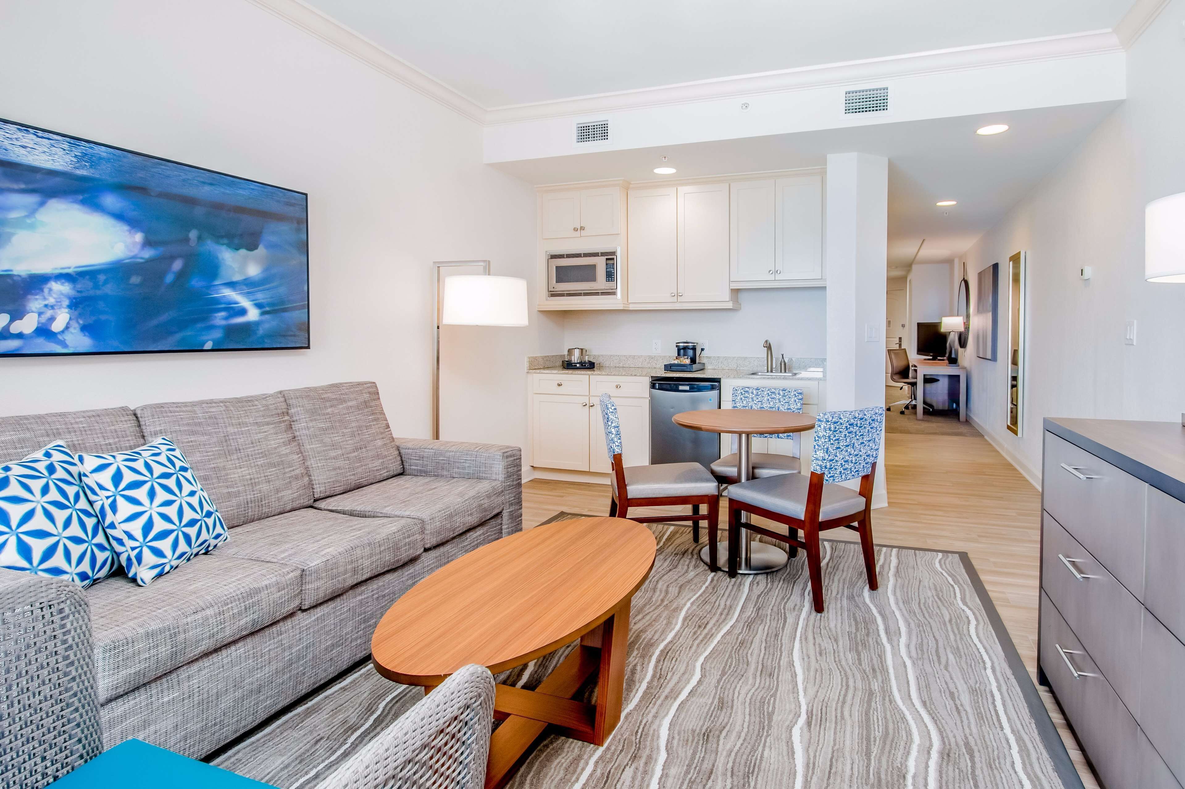 A modern living room with a sofa, coffee table, and chairs, a kitchen area with cabinets and appliances, and a dining table with chairs.