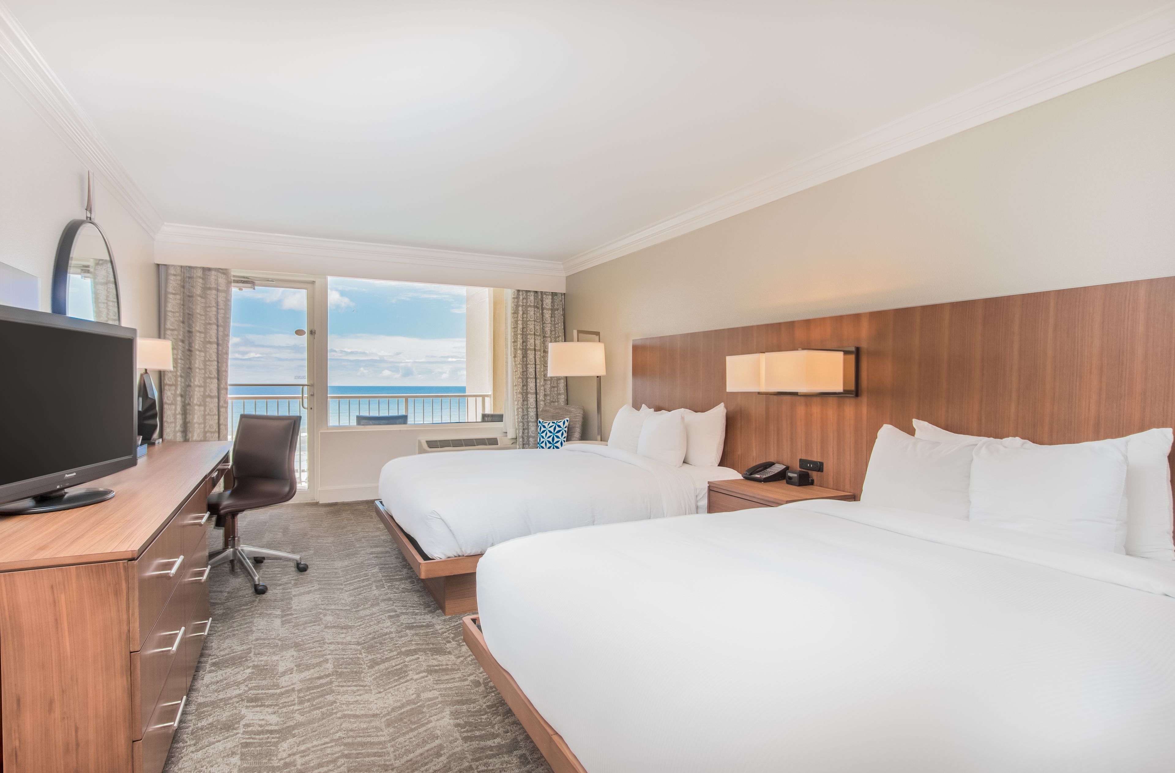 A hotel room with two beds, a desk with a TV, and a view of the ocean.