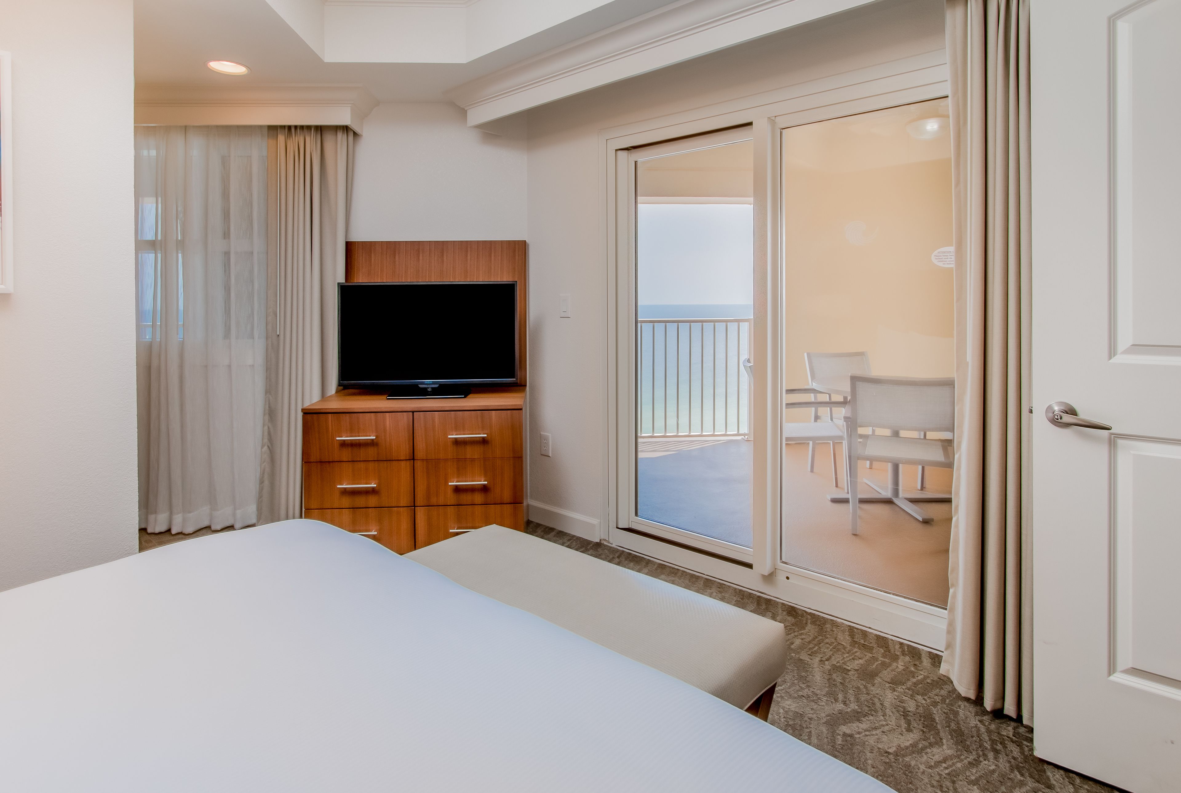 Une chambre d’hôtel avec un lit, une télévision, une commode en bois et un balcon avec des chaises.