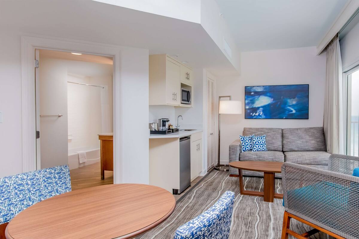 Une chambre d’hôtel avec une kitchenette, un coin repas doté d’une table ronde, un canapé et un grand tableau abstrait.