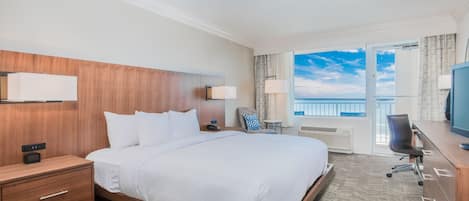 A hotel room with a large bed, bedside tables, a desk with a chair, a television, and a view of the ocean through a balcony door.