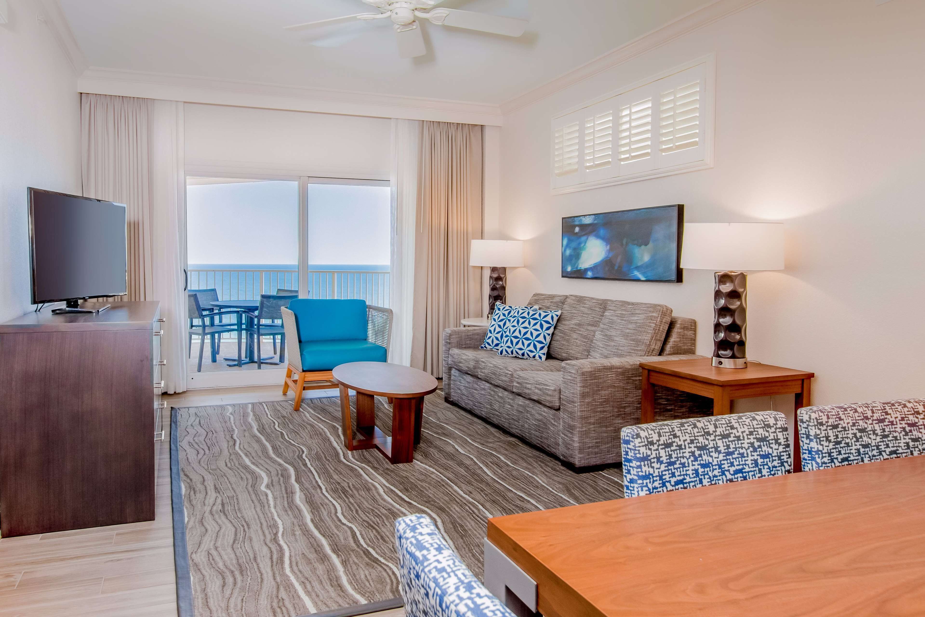 A hotel room with a balcony, a TV, a sofa, a table, and a patterned rug.