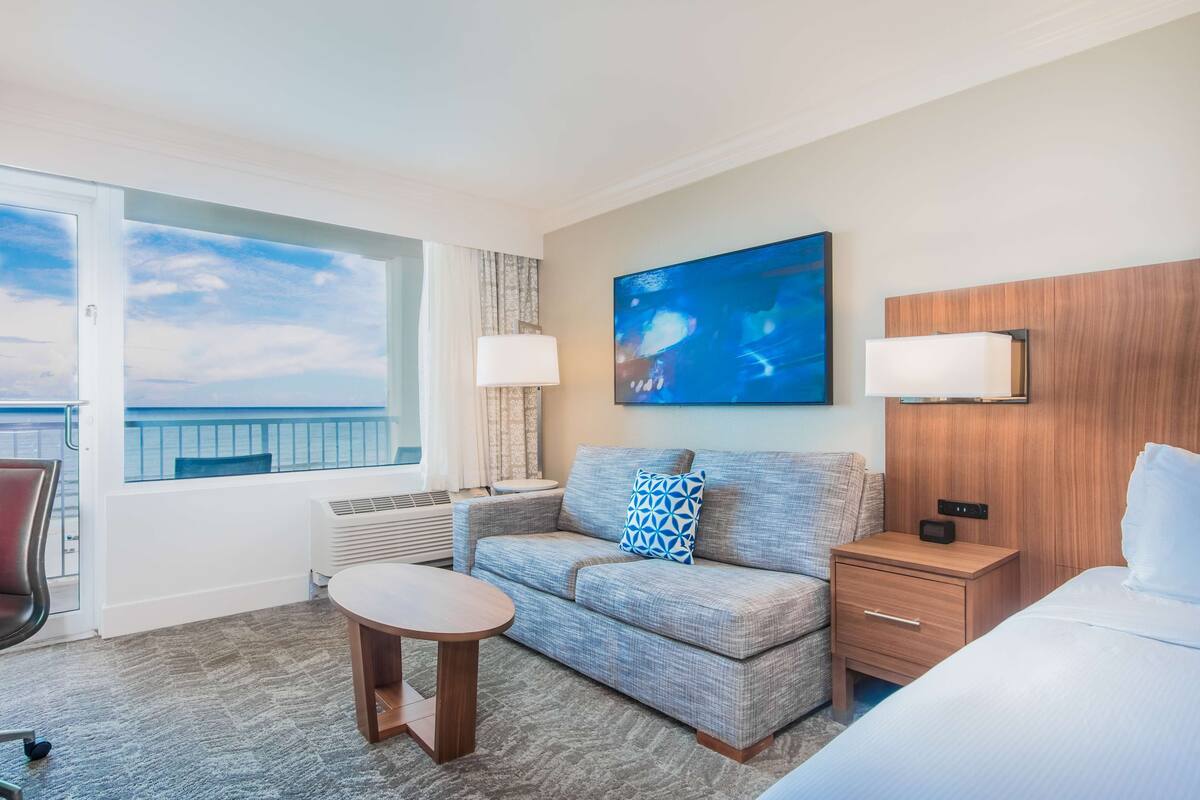 Une chambre d’hôtel avec une grande fenêtre, un canapé, une table de chevet, une lampe et un lit.