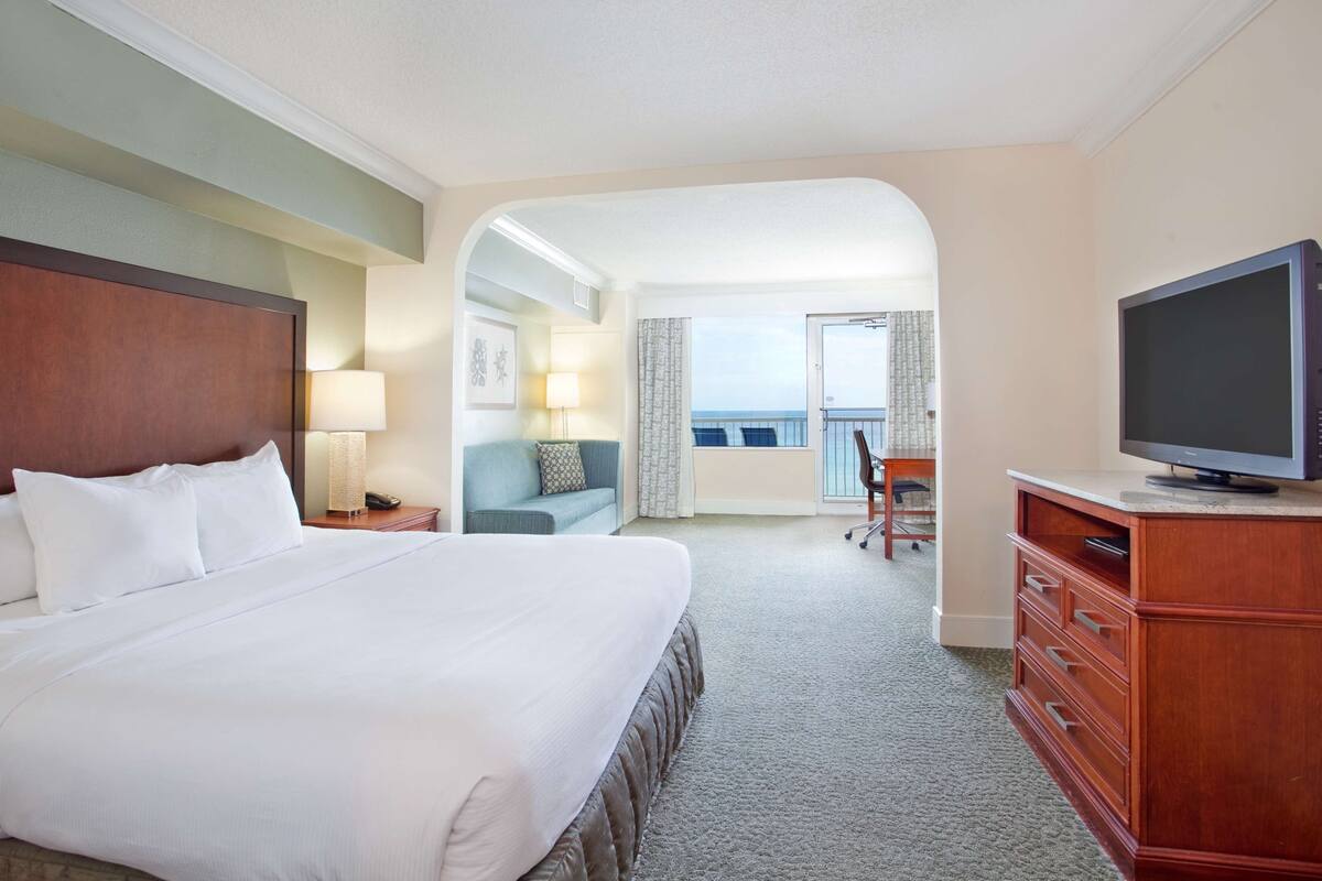 Une chambre d’hôtel avec un grand lit, un téléviseur posé sur une commode en bois, un balcon avec vue et un coin salon comprenant un canapé et un fauteuil.
