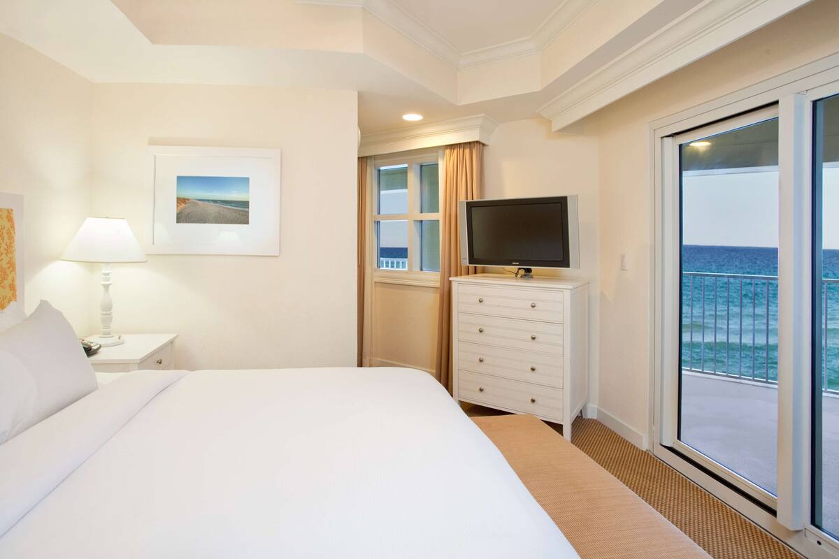 Une chambre d’hôtel avec un grand lit, un téléviseur posé sur une commode, une fenêtre donnant sur la mer et un tableau encadré au mur.