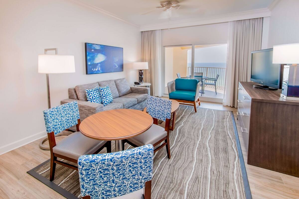 Une chambre d’hôtel moderne avec un canapé, une table ronde et des chaises assorties. On y trouve également une télévision et un bureau avec une lampe. Un tableau est accroché au mur, et on aperçoit un balcon à travers la fenêtre.