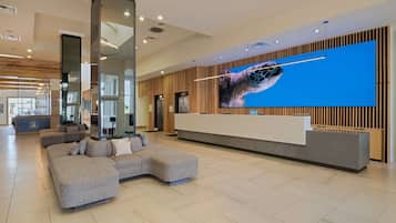 A modern hotel lobby with a reception desk, a large digital screen displaying a sea turtle, and a seating area with a sectional sofa.