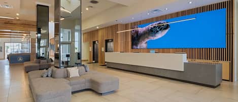 A modern hotel lobby with a reception desk, a large digital screen displaying a sea turtle, and a seating area with a sectional sofa.