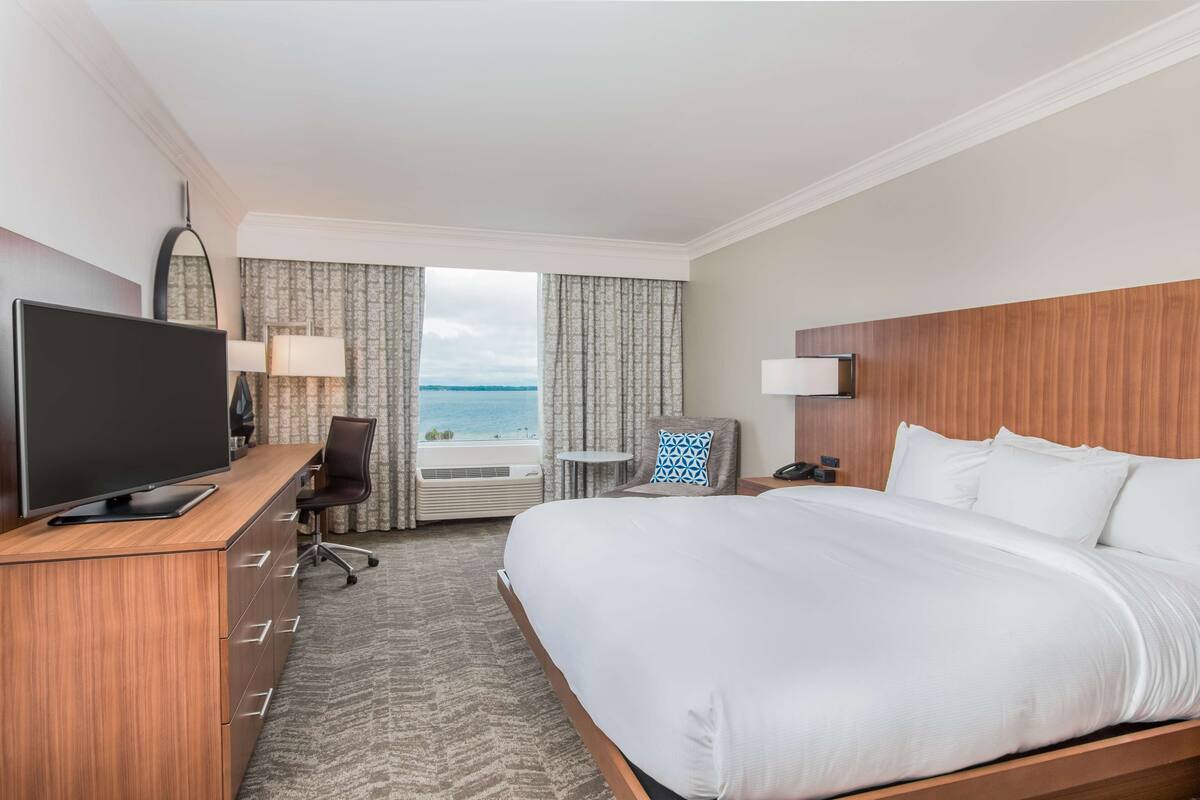 Une chambre d’hôtel avec un grand lit, un bureau avec une télévision, une chaise et une vue sur l’océan par la fenêtre.