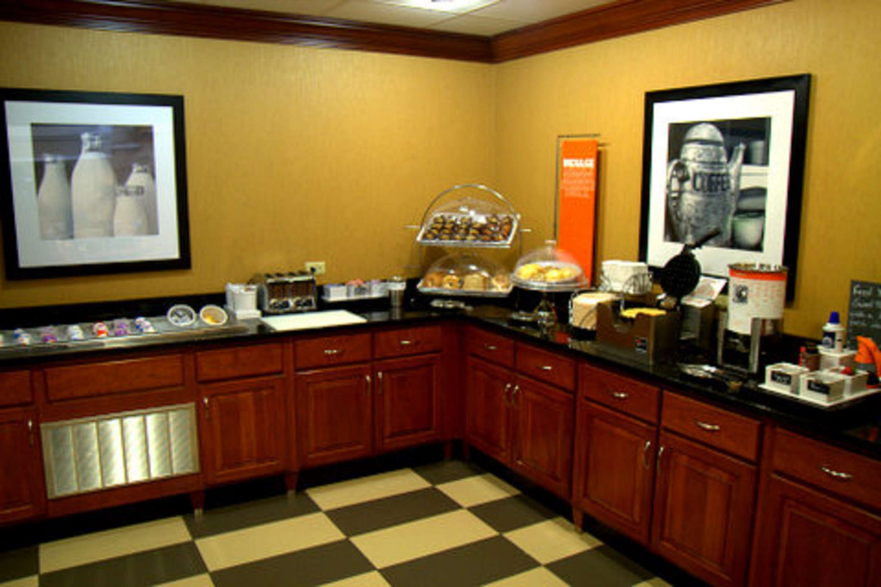 A hotel kitchen with wooden cabinets, a checkered floor, a coffee station, and framed pictures on the walls.
