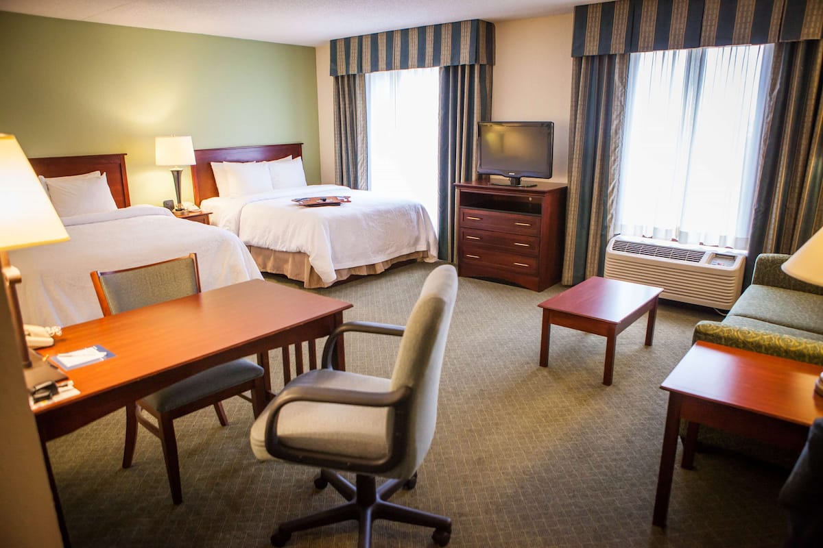 A hotel room with two beds, a desk, a chair, a television, and an air conditioning unit.