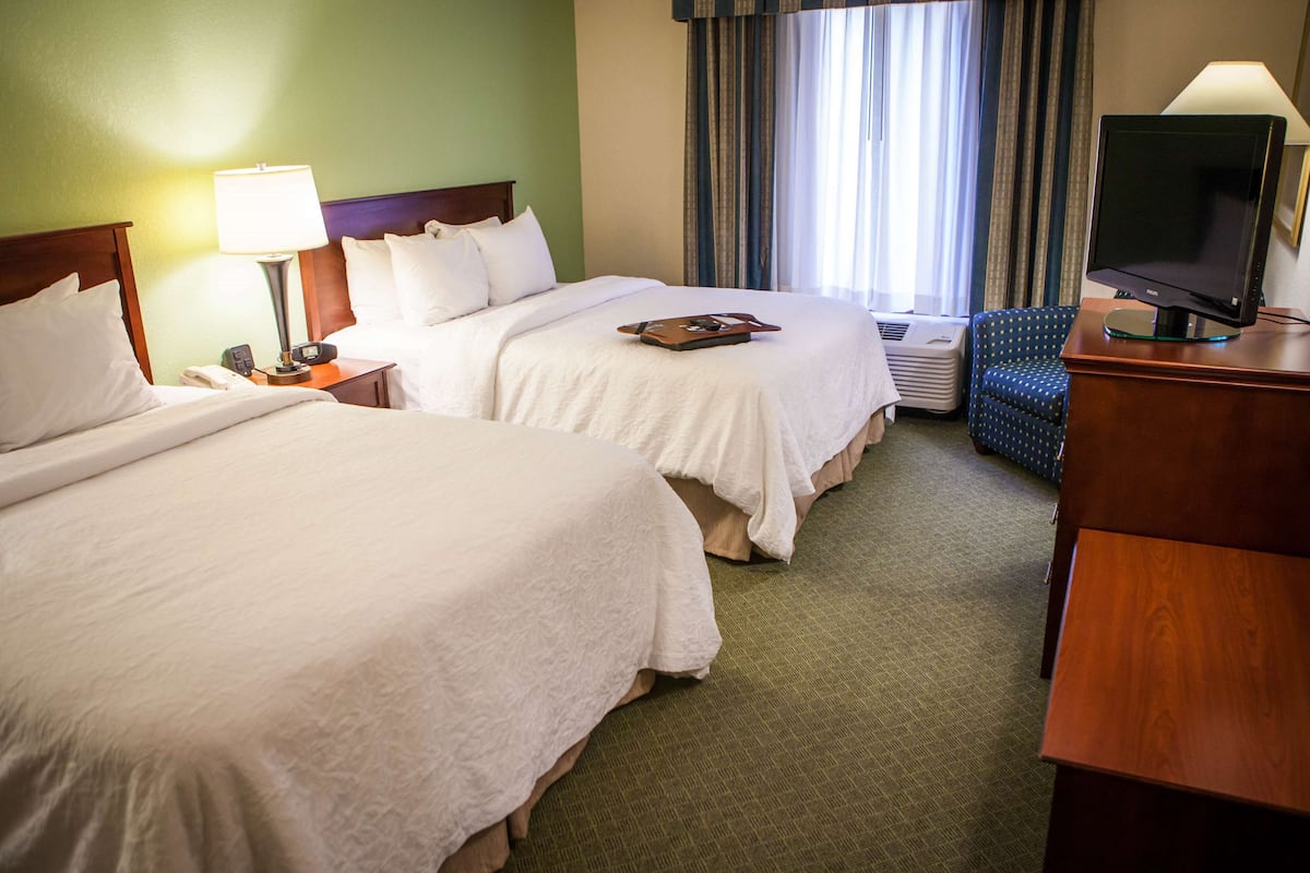 A hotel room with two beds, a desk with a television, a chair, and a window with curtains.