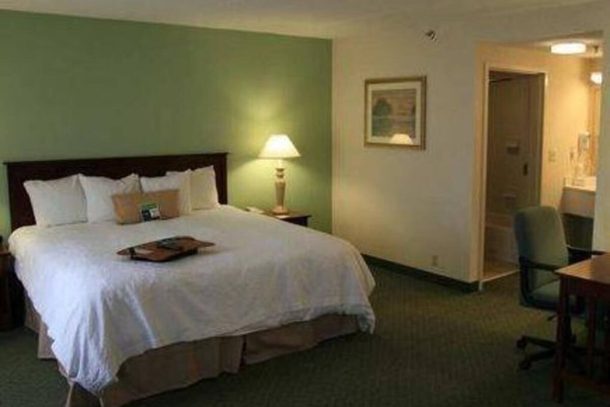 A hotel room with a bed, a desk, a chair, a lamp, and a painting on the wall.