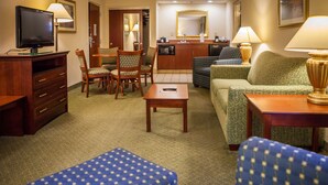Quarto de hotel com televisão, mesa de jantar com cadeiras, sofá, mesa de centro e cozinha compacta.