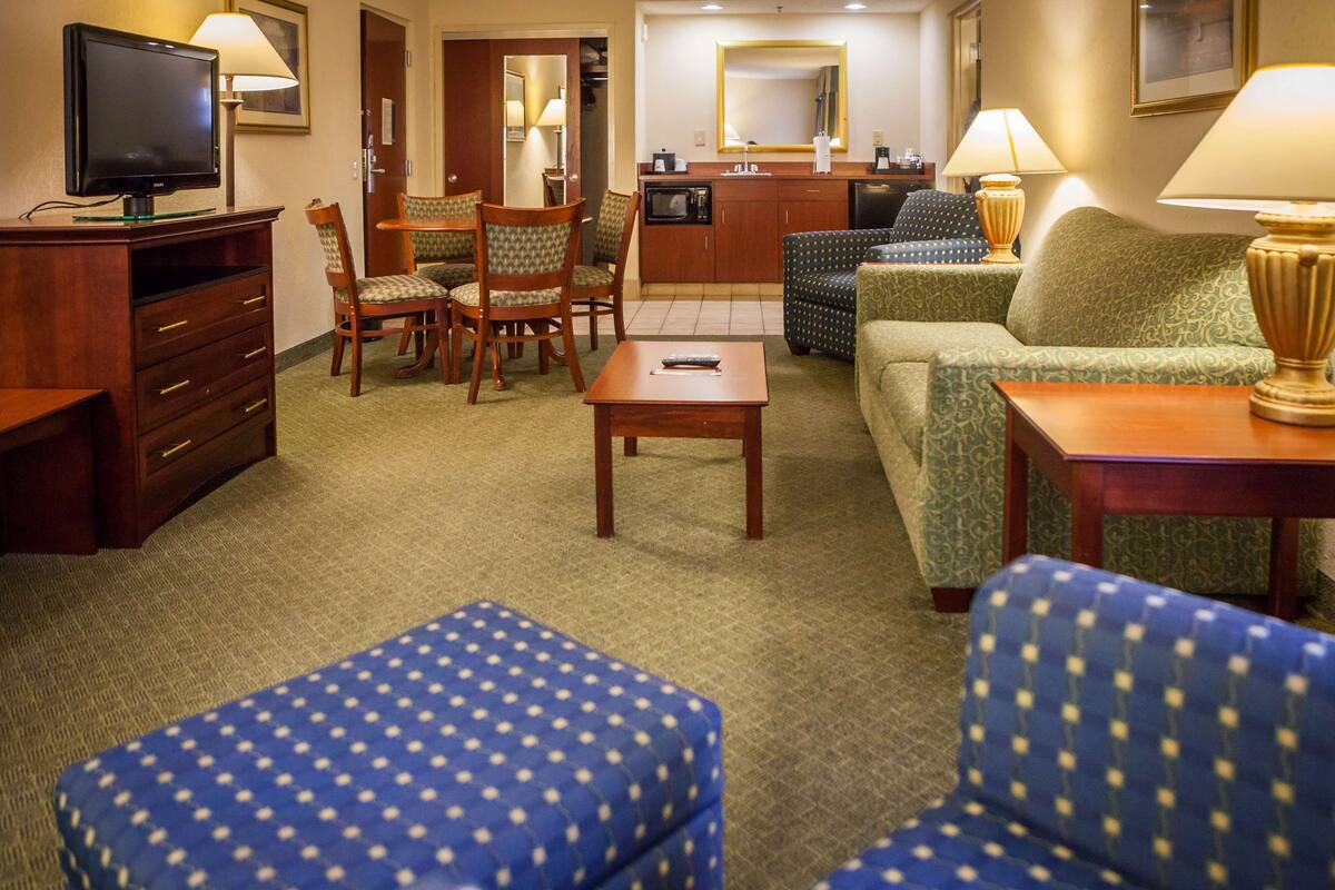 A hotel room with a TV, a dining table with chairs, a sofa, a coffee table, and a kitchenette area.