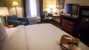 A hotel room with a large bed, a desk with a computer, a chair, a TV, and a minibar.