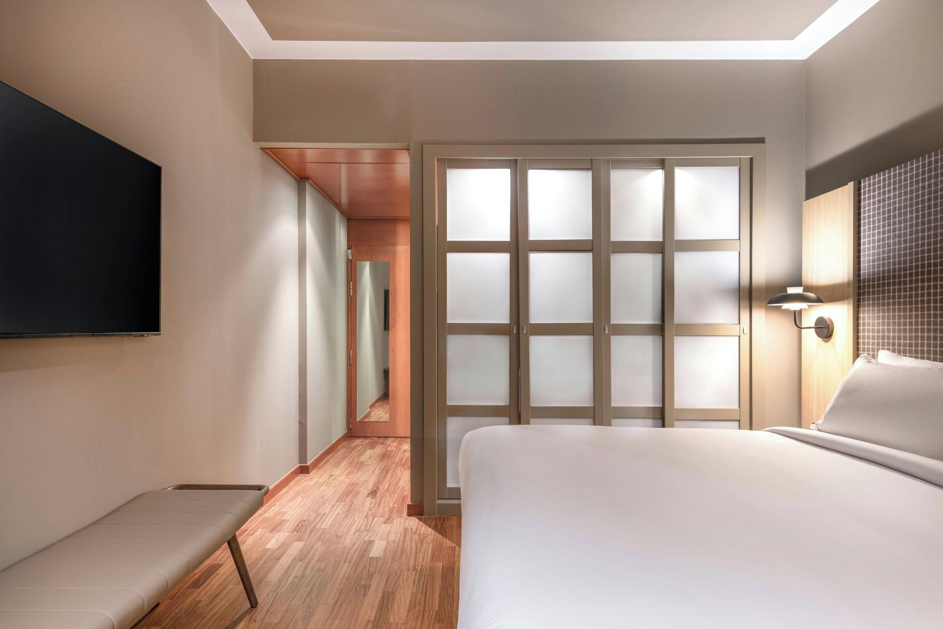 Une chambre d’hôtel comprenant un lit, une banquette, un téléviseur, une porte coulissante et une lampe fixée au mur.