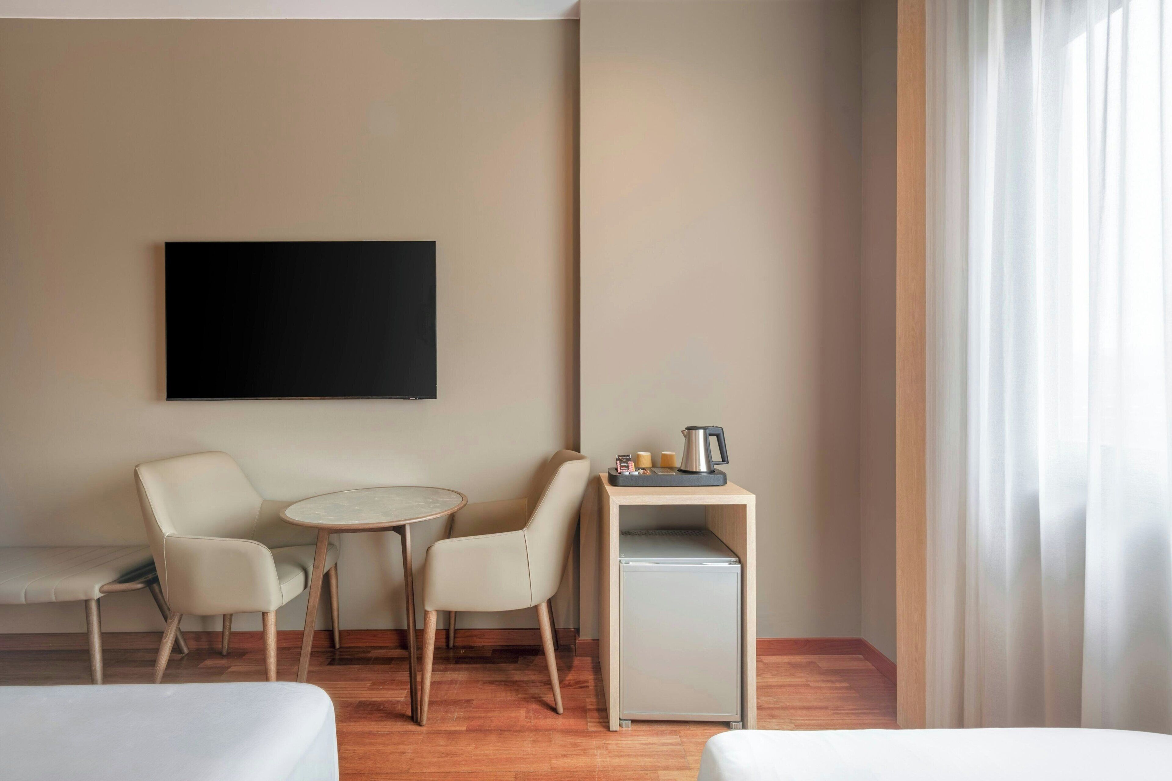 Habitación de hotel con cama, dos sillas, una mesita, cafetera y televisor montado en la pared.