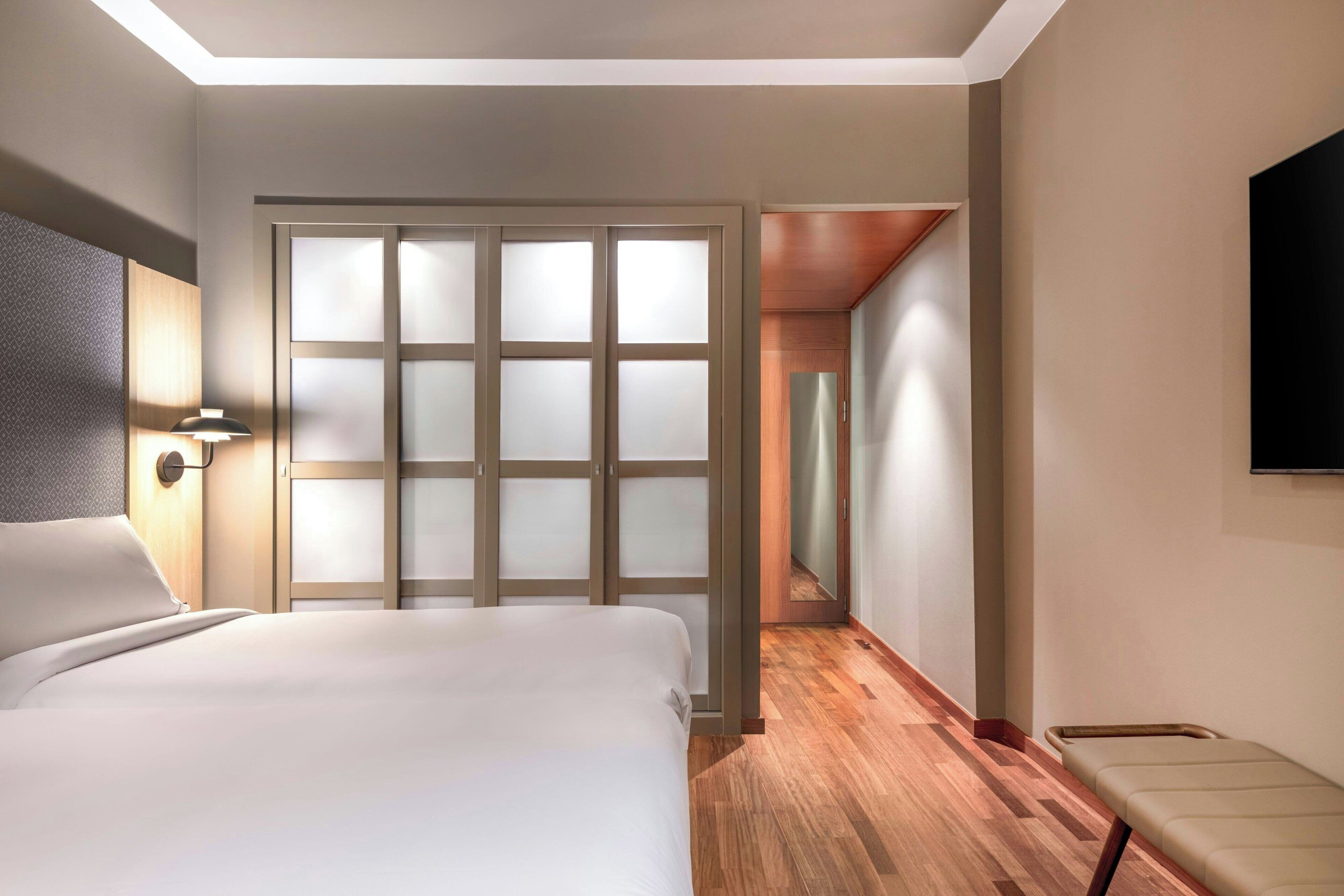 A hotel room with a bed, a wall-mounted TV, a wooden floor, and a sliding door with frosted glass panels.