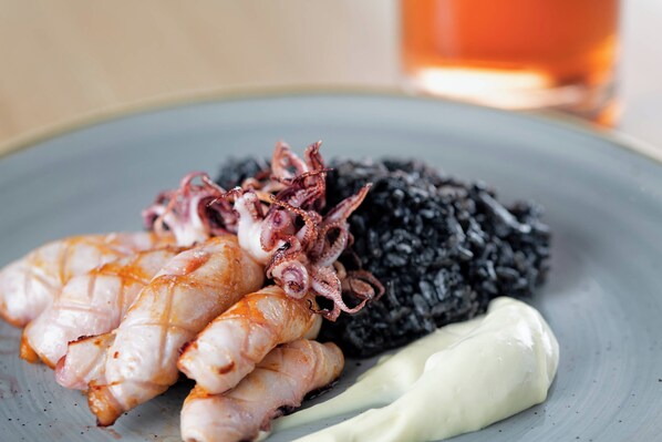 Un plato con pulpo a la parrilla, calamares y arroz negro, acompañado de salsa blanca.