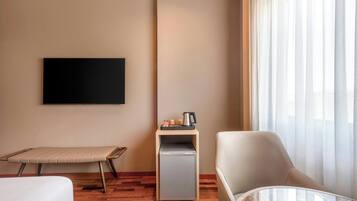 Habitación de hotel con cama, zona de estar con una silla y una mesa, kitchenette pequeña, televisor de pantalla plana montado en la pared y ventana con cortinas translúcidas.