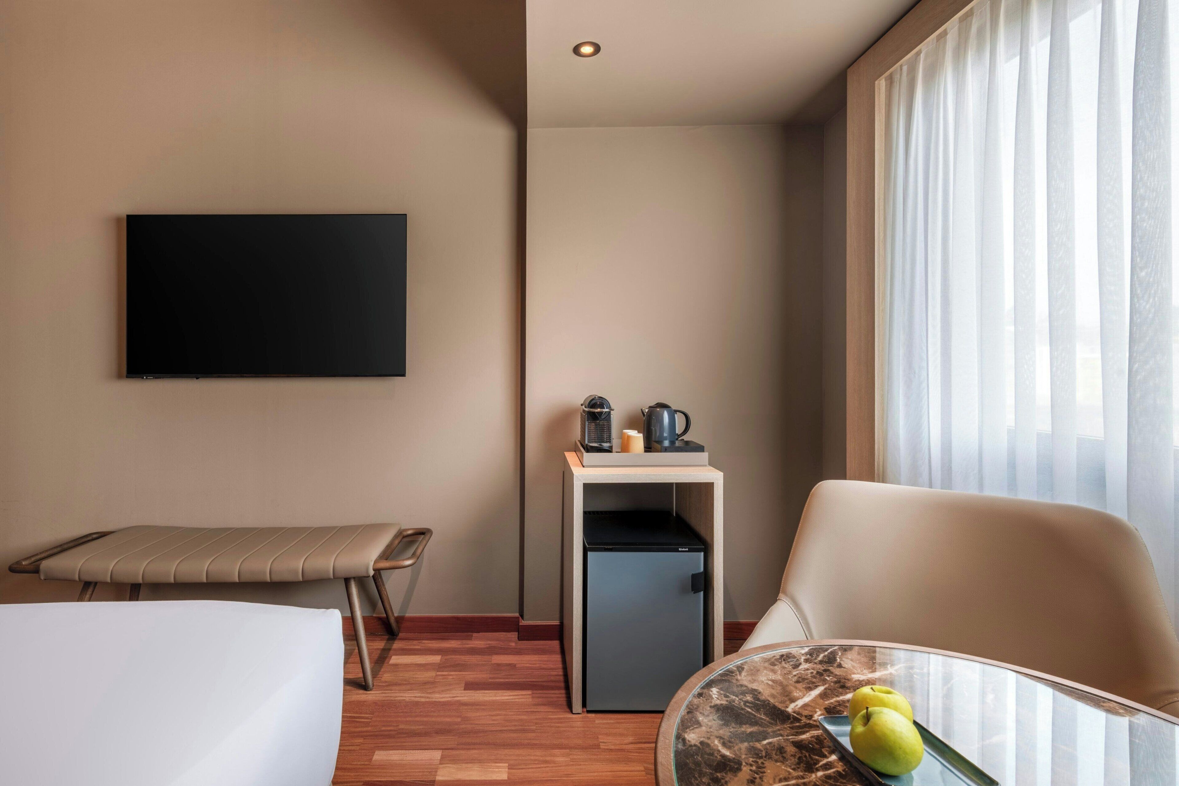 A hotel room with a bed, a desk with a computer, a chair, a small refrigerator, and a television mounted on the wall.