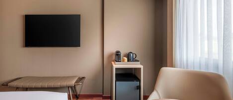 A hotel room with a bed, a desk with a computer, a chair, a small refrigerator, and a television mounted on the wall.