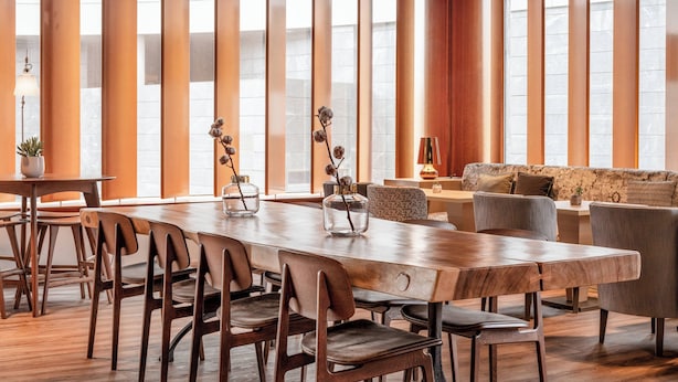 Un comedor amplio con una larga mesa de madera y sillas, ventanas grandes con persianas verticales y un rincón de estar acogedor con un sofá y sillones.