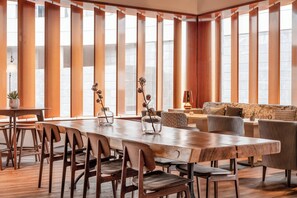 Un comedor amplio con una larga mesa de madera y sillas, ventanas grandes con persianas verticales y un rincón de estar acogedor con un sofá y sillones.