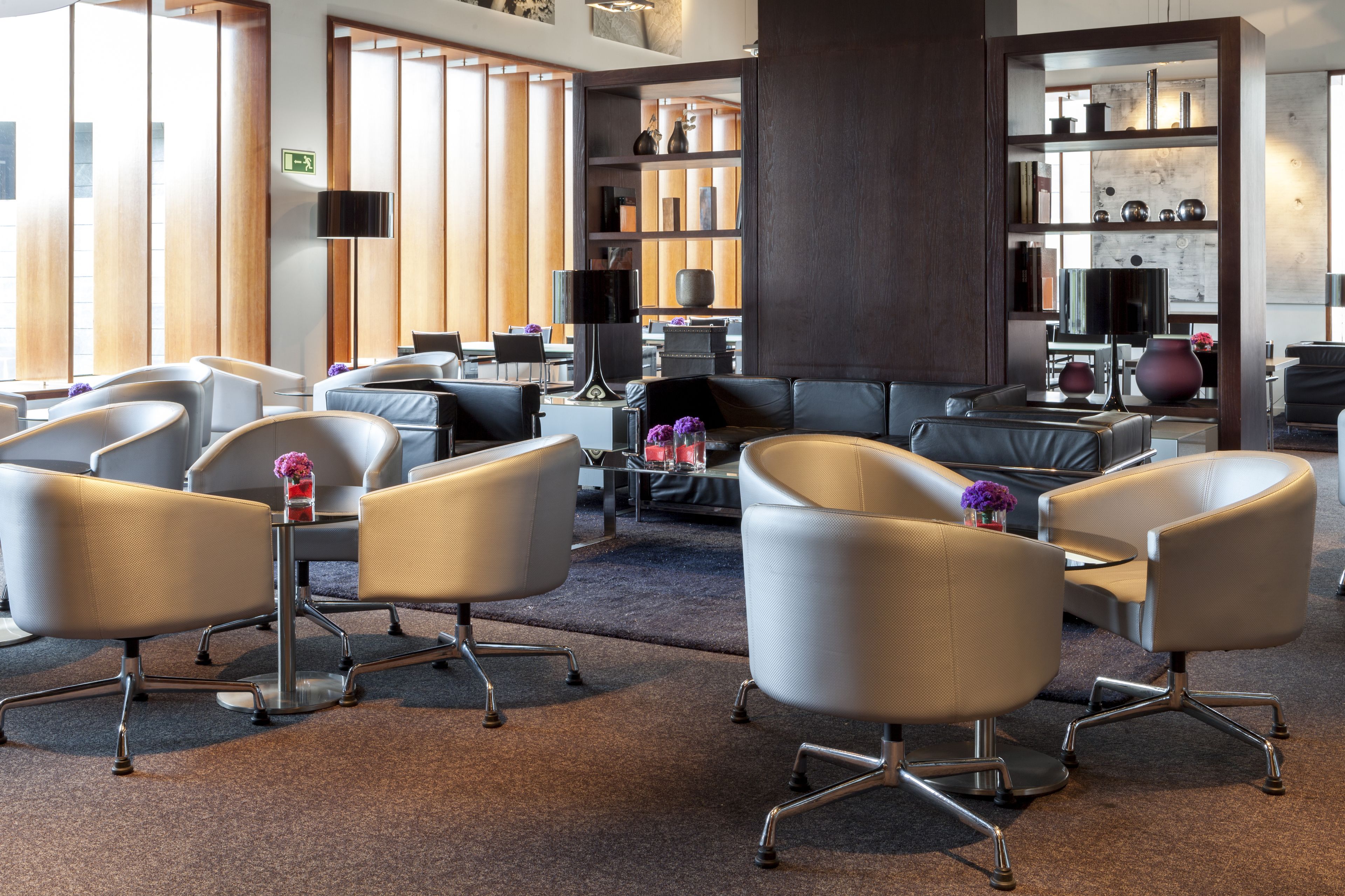 A modern lounge area with leather sofas, armchairs, and a coffee table. There is a bookshelf with decorative items and a flat-screen TV mounted on the wall.