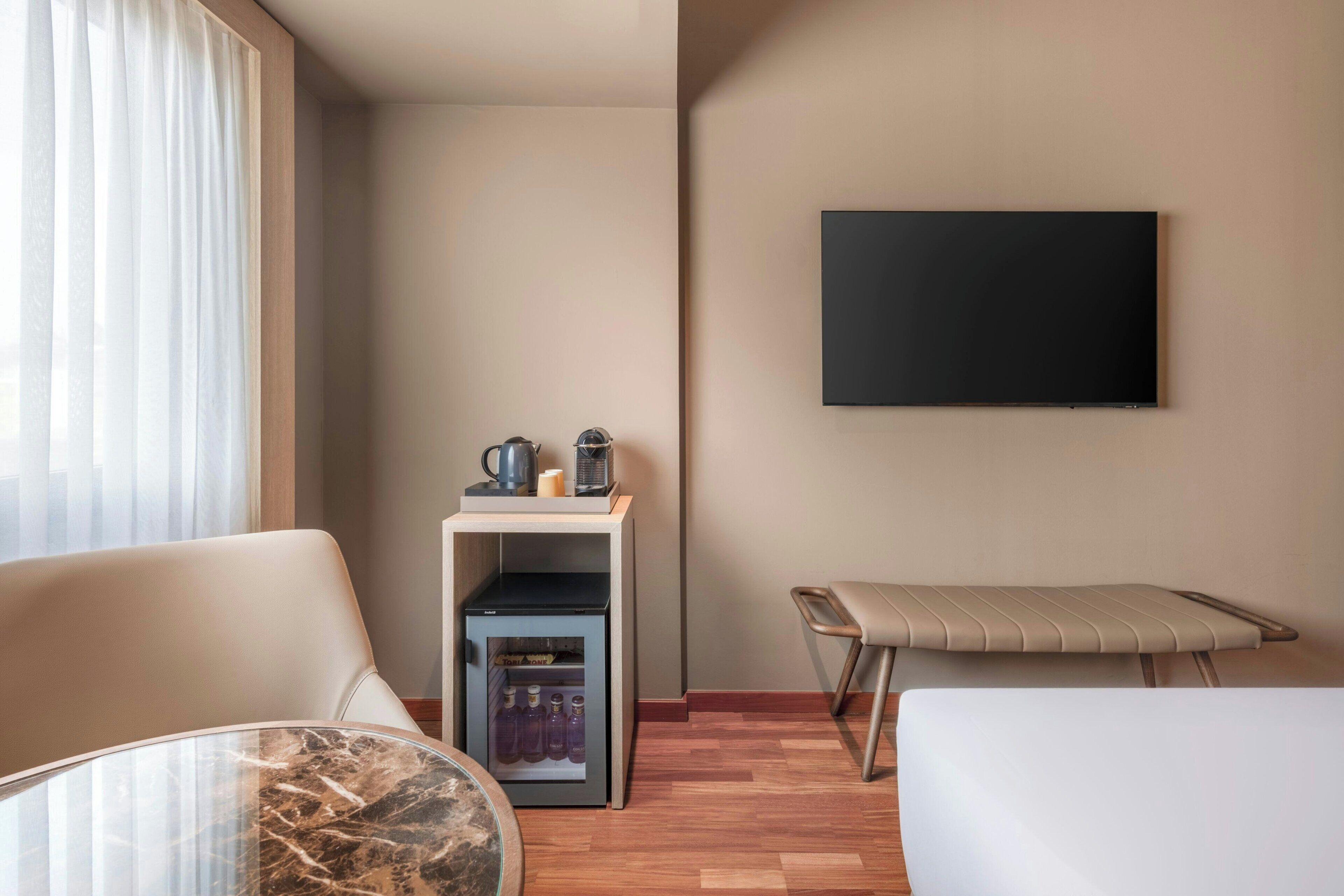 A hotel room with a bed, a seating area with a table, a small kitchenette, a TV mounted on the wall, and a window with curtains.